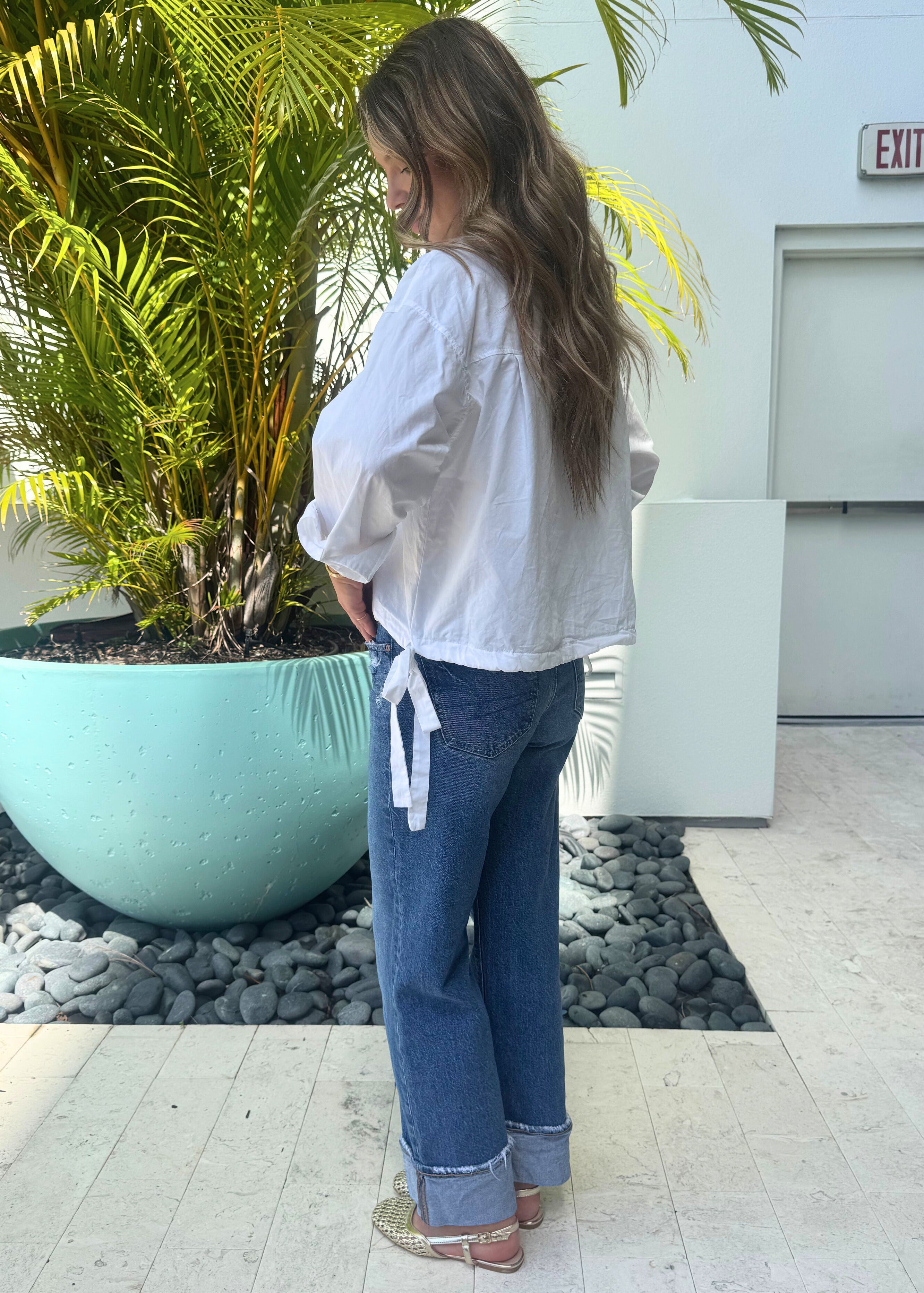 Woman standing outdoors next to a large planter with palm trees, wearing a white blouse and blue jeans.