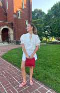 Woman in a white outfit with a red handbag standing on a brick path with green grass and trees in the background.