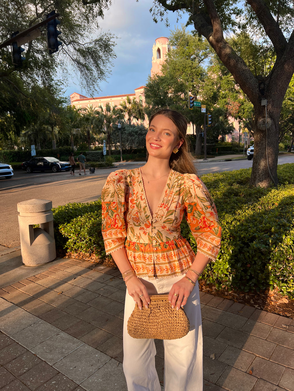 Woman in a colorful peplum top holding a woven bag on a city street with trees and buildings in the background.