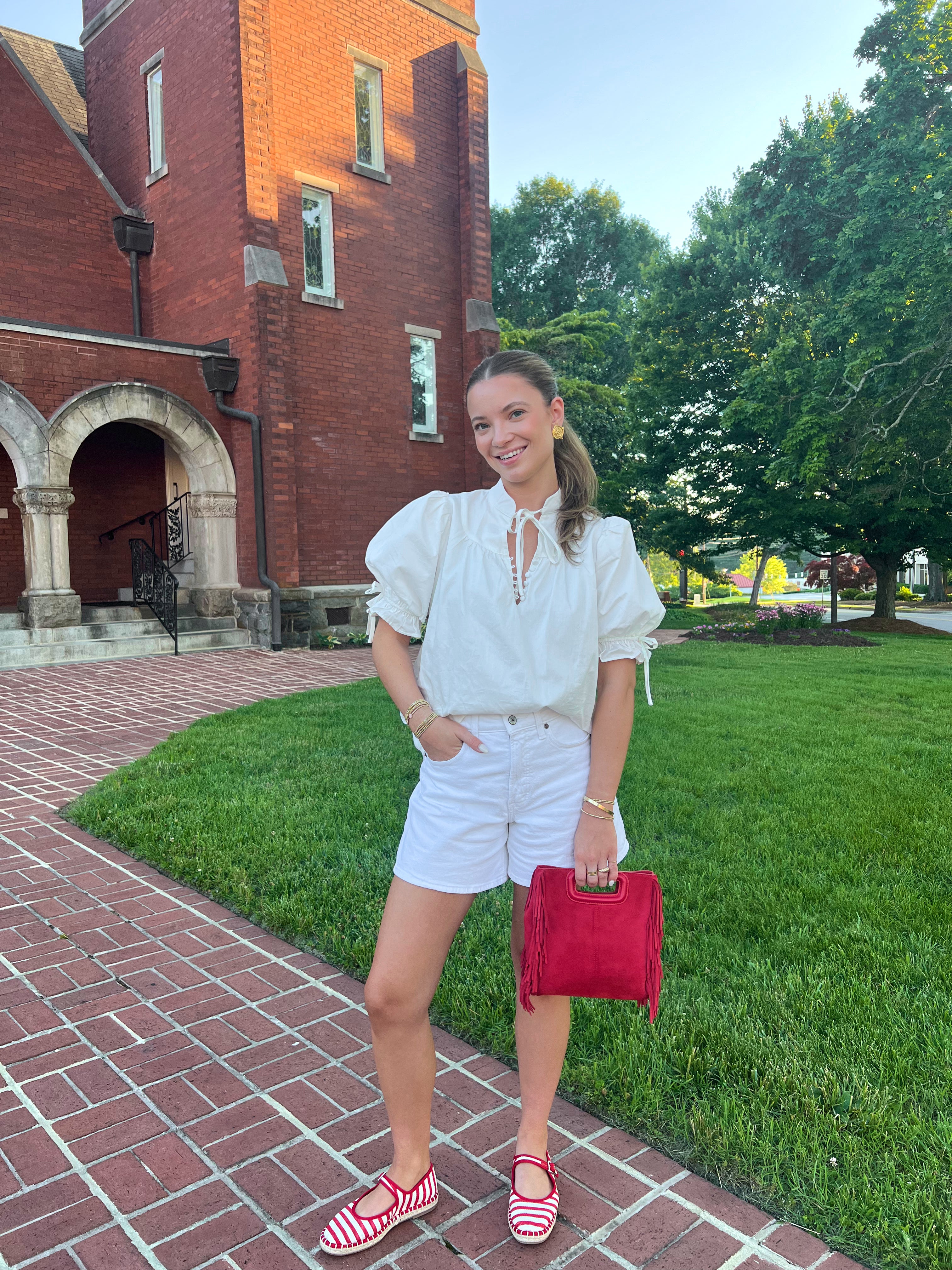 Woman in white outfit with red clutch standing on a brick path in front of a brick building.