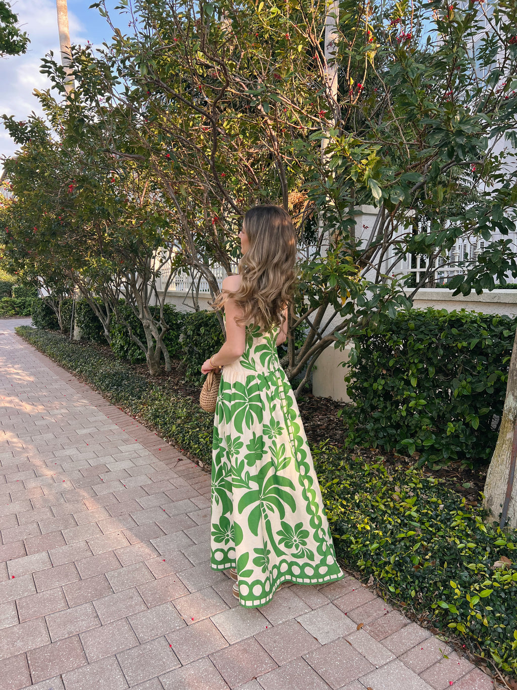 Woman in a green and white dress standing on a sidewalk with trees and bushes in the background