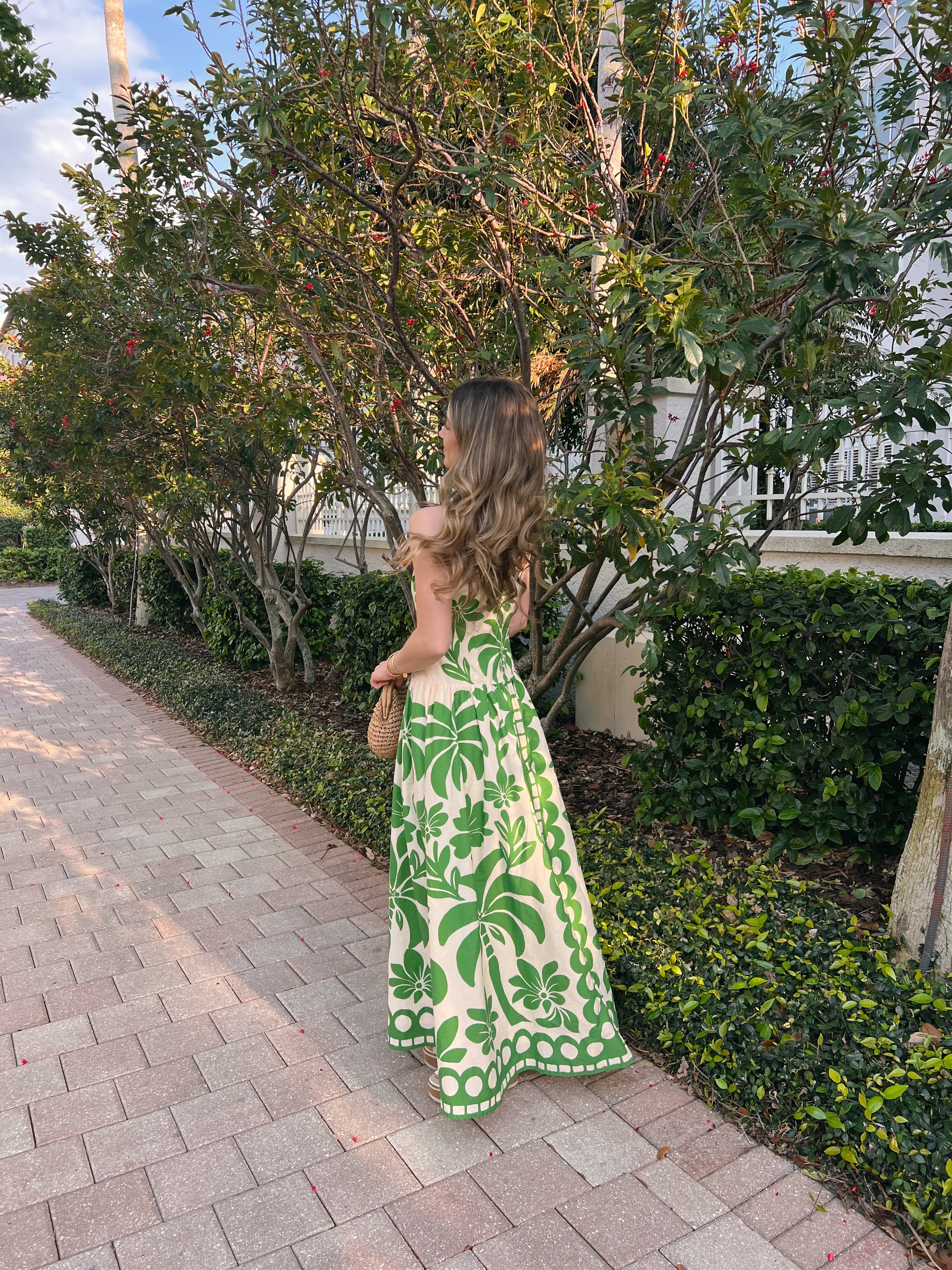 Woman in a green and white dress standing on a sidewalk with trees and bushes in the background