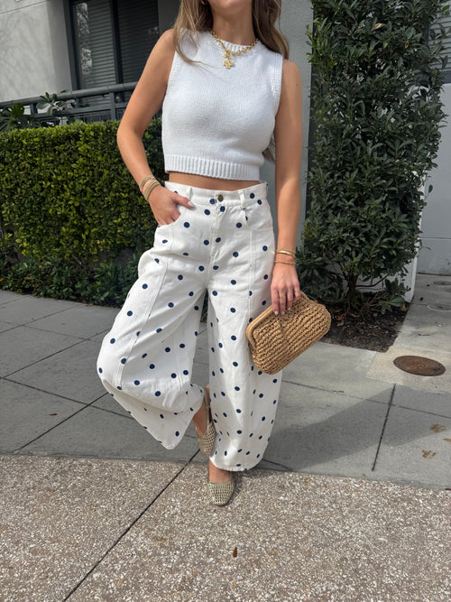 Person wearing a white outfit with black polka dots, holding a woven bag on a sidewalk.