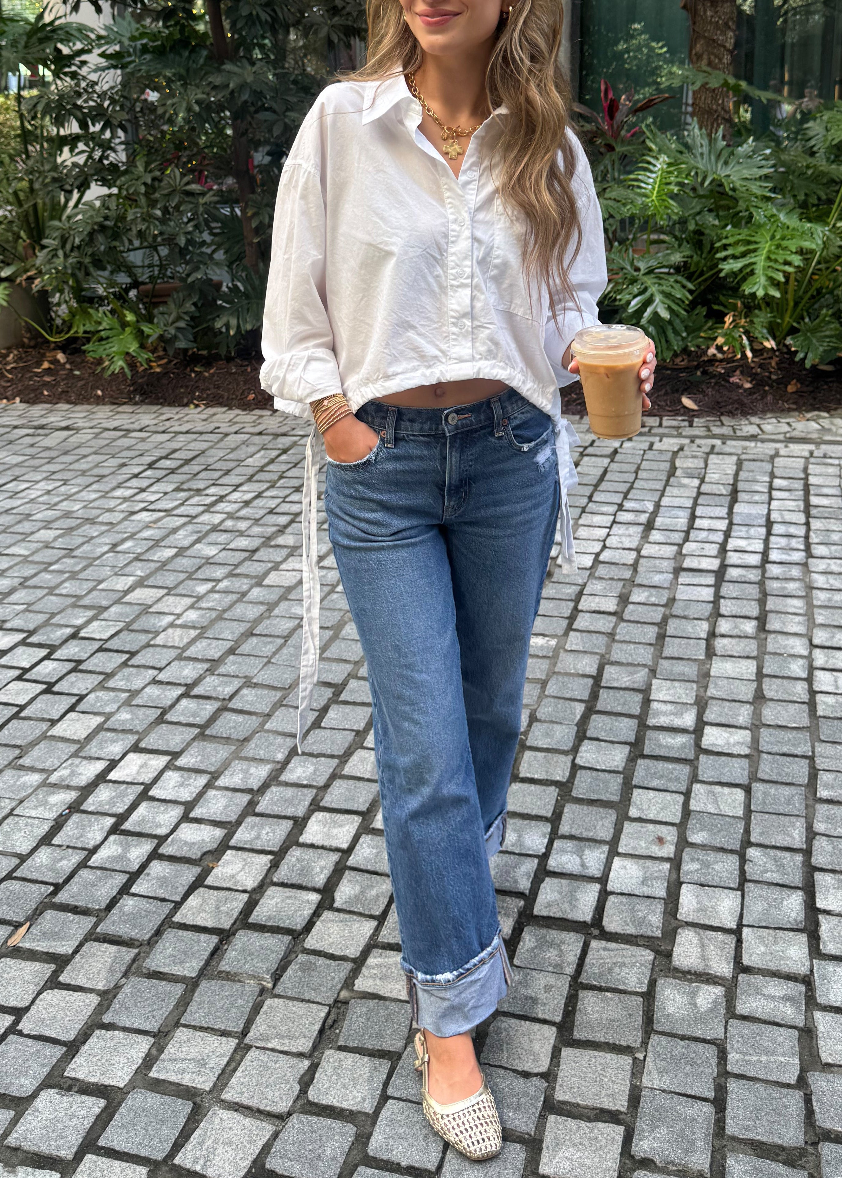 Person wearing a white shirt and blue jeans standing on a paved path with greenery.
